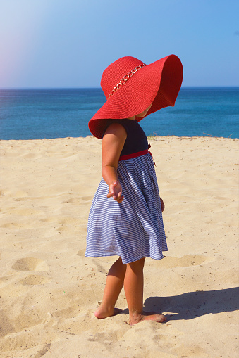 A little girl in a red hat and a blue dress walks on the beach on the sand by the sea. High quality photo