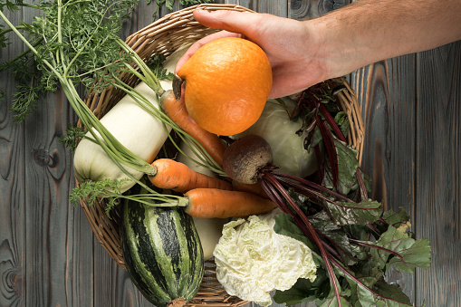 Unrecognizable cropped man hand holding little orange pumpkin against woven basket of tasty, juicy raw vegetables on wooden table. Raw food unpack, delivery, donation and charity relief. Top view