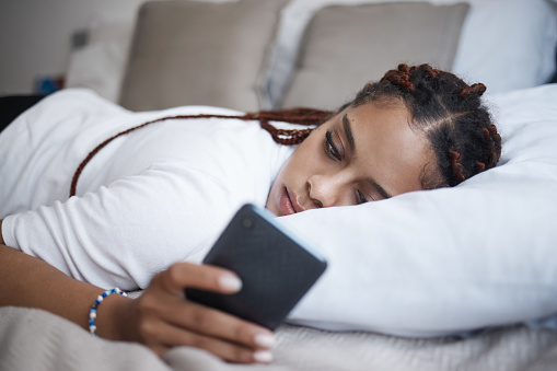 Depresión, mujer triste y africana en un teléfono relajándose en la cama de su habitación en casa. Cansada, de salud mental y chica negra solitaria navegando en las redes sociales o Internet mientras tiene problemas de ruptura. photo