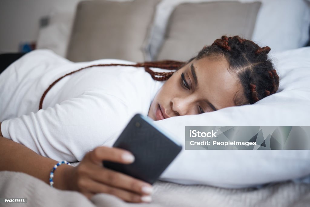 Depression, sad and african woman on a phone relaxing on the bed in her bedroom at home. Tired, mental health and lonely black girl browsing on social media or internet while having breakup problems. Depression - Sadness Stock Photo