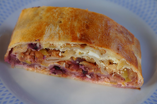 The piece of home made apple strudel on plate, stuffed with grated apples, butter, cheese cream  and blueberry, on rolled dough is in baking pan with baking paper. Butter and egg spread is added to the apple strudel.