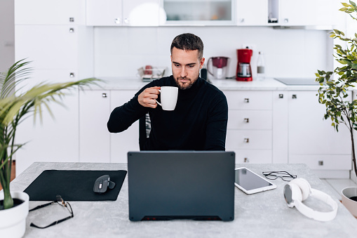 A young Caucasian man is holding a cup of coffee while sitting in front of his laptop and working from home.