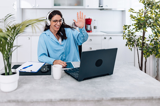 A young Caucasian woman is waving while having a video conference with her colleagues. She's working from her home office.