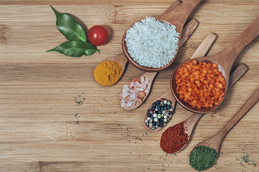 Spices on wooden spoon on wooden background. Big spoons with white rice and red lentil, small spoons with rock salt, black pepper, red pepper, dill and turmeric, decorated with cherry tomatoes and fresh Bay leaf. Horizontal photo.