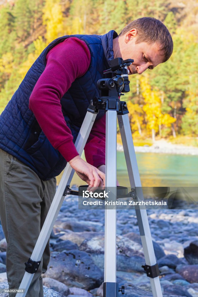 Young attractive man sets up metal tripod outdoors to observe celestial bodies through telescope. Astronomy work.Autumn day, travel, hobby and DIY, on bank of mountain river, tourist sets up tripod for photo, video camera or telescope.Selective focus, side view. 40-44 Years Stock Photo
