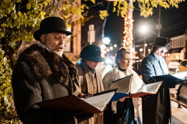 Mature people singing Christmas carols outdoors at night.  They are dressed up in period costumes. Exterior of city street with Christmas decor.