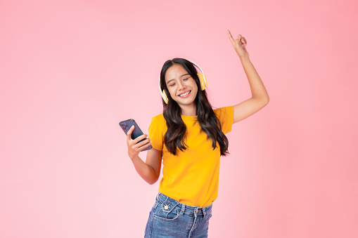 Cheerful young Asian woman in headphones listening to music and enjoy favourites playlist application on smartphone with dancing on pink background.