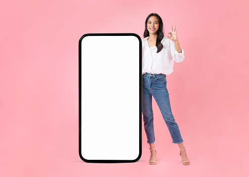 Beautiful Asian woman shows ok sign and stand with big smartphone mockup of blank screen and smiling on pink background.