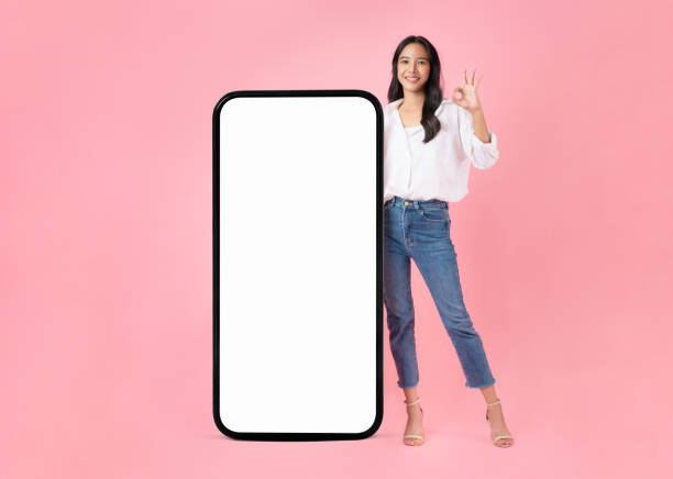 Beautiful Asian woman shows ok sign and stand with big smartphone mockup of blank screen and smiling on pink background. stock photo