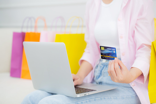 Young asian woman sitting on sofa using laptop computer shopping online with credit card while paper bag on sofa, female paying with transaction financial, purchase and payment, business concept.