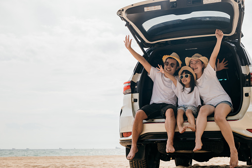 Happy Family Day. Dad, mom and daughter enjoying road trip sit on back car and raise hand up, Family traveling in holiday at sea beach, people having fun in summer vacation on beach with automobile