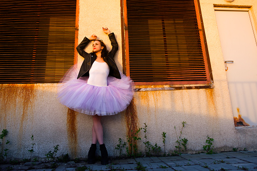 a ballerina in a tutu and a black jacket stands against the wall of an old house pretending to take off