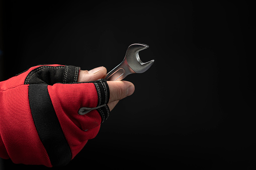 A strong male hand in a red glove holds a wrench on a black background. The concept of plumbing, mechanical and automotive work.