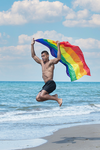 Portrait of a man holding and waving a rainbow gay pride flag while jumping on the beach.