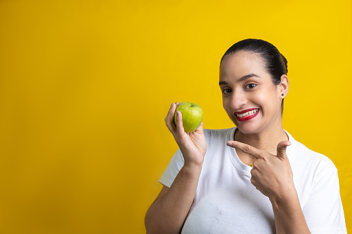 A funny Hispanic woman pointing to a green apple isolated on a yellow background