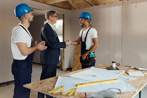 Happy real estate developer shaking hands with one worker at construction site while the other worker is applauding. Copy space.