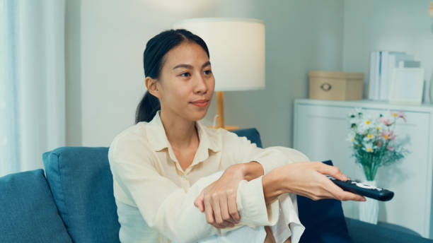 istock Beautiful Asian young woman on sofa click remote controller change channel on television choose favorite comedy or sitcoms in streaming online VOD, video on demand at home. 1443178878