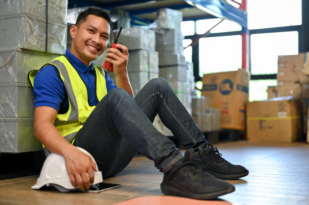 istock Happy Asian male warehouse worker in uniform sitting in warehouse, using walkie-talkie. 1443178921
