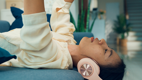 Close-up Young Asia university girl lay down on sofa reading textbook wear headphone listen aural, auditory learning on audio streaming broadcast online at home. Leisure activity, Lifestyle concept.