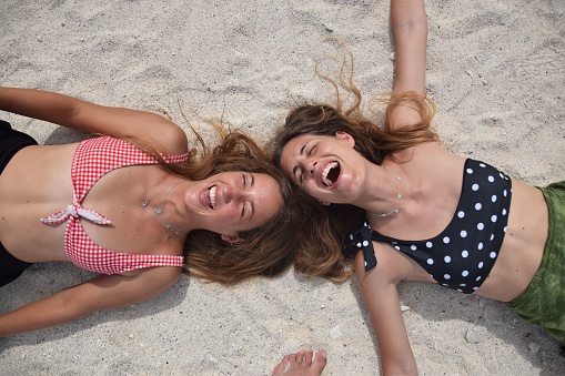A top view of two happy young caucasian women in swimsuits lying on the sandy beach and laughing