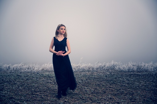 A young Caucasian female in a long black dress standing in a field with foggy background
