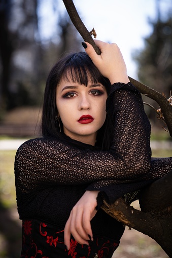 A Portrait of a pretty female in a gothic style with a red lipstick looking at camera in the park