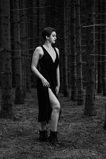 of A grayscale Young Caucasian female in a long black dress standing in the forest