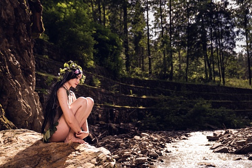 a Pretty caucasian girl in brown short dress with a flowers crown sitting on a rock in the park river