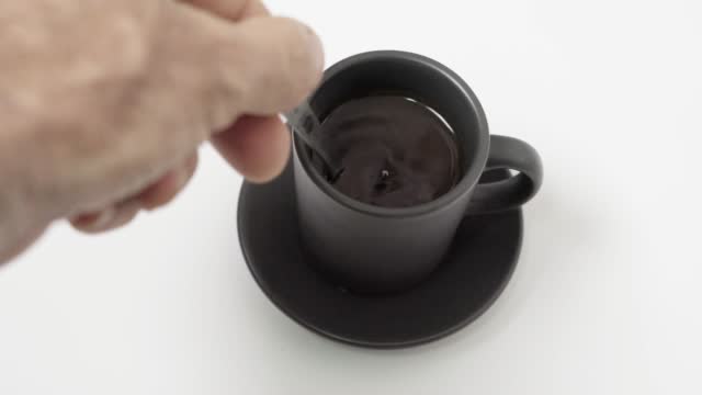 4K video of a person using a spoon to stir black coffee in a small black mug on a white surface