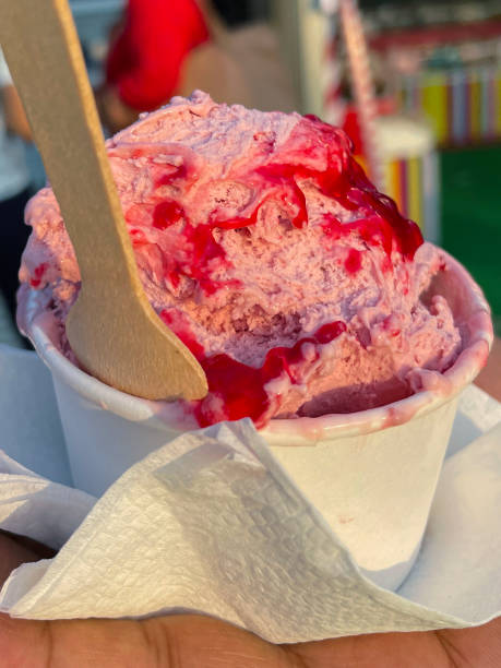 istock Full frame image of white, single-use, disposable, cardboard tub of strawberry and raspberry gelato with berry fruit syrup held on unrecognisable person's palm, wooden spoon, paper napkin, focus on foreground 1443190689