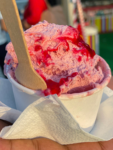 Stock photo showing a close-up view of street food dessert of scoops of Summer fruit gelato and red berry syrup served in a white, single-use, disposable, cardboard tub with wooden spoon.