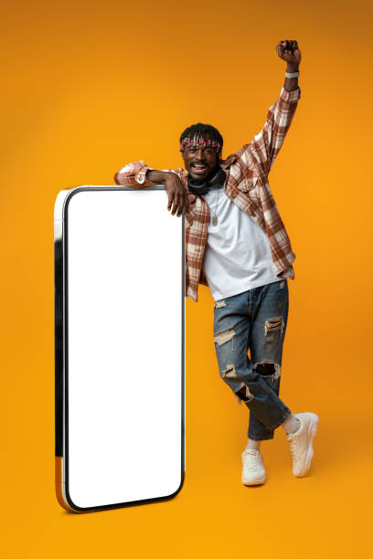 istock Smiling african man standing near huge smartphone with blank screen on yellow studio background 1443191705