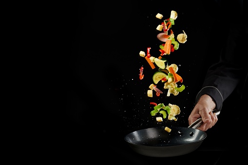 Crop unrecognizable male chef in uniform throwing fresh delicious assorted vegetables while preparing healthy dinner in frying pan against black background