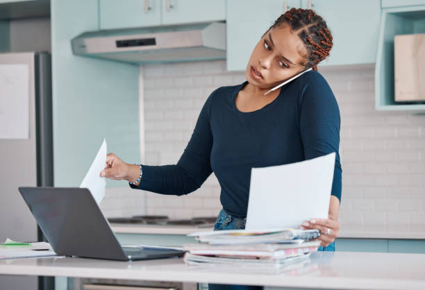 donna d'affari nera che lavora da casa in scartoffie freelance, multitasking con laptop e telefonata audio mobile. tecnologia internet in cucina, consultazione dei documenti di fatturazione nella pandemia covid - economic crisis audio foto e immagini stock