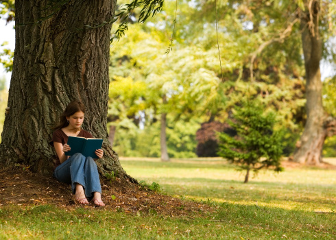 A middle-school girl readying in the shade of a tree in a park-like setting.