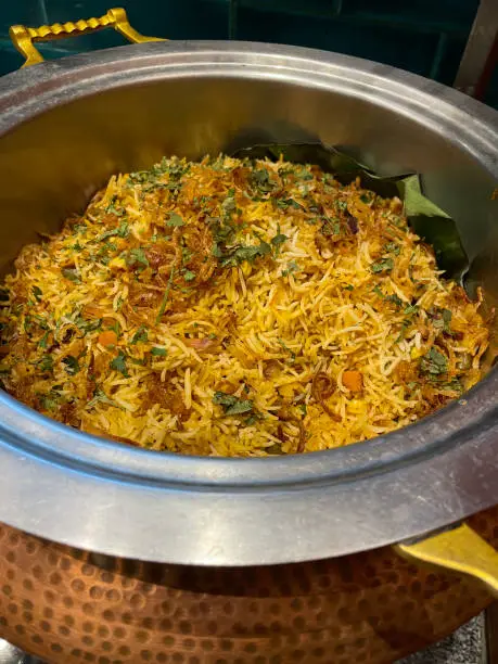 Stock photo showing close-up, elevated view of rice dish of chicken biryani in dimpled, copper metal catering dish on a restaurant counter.