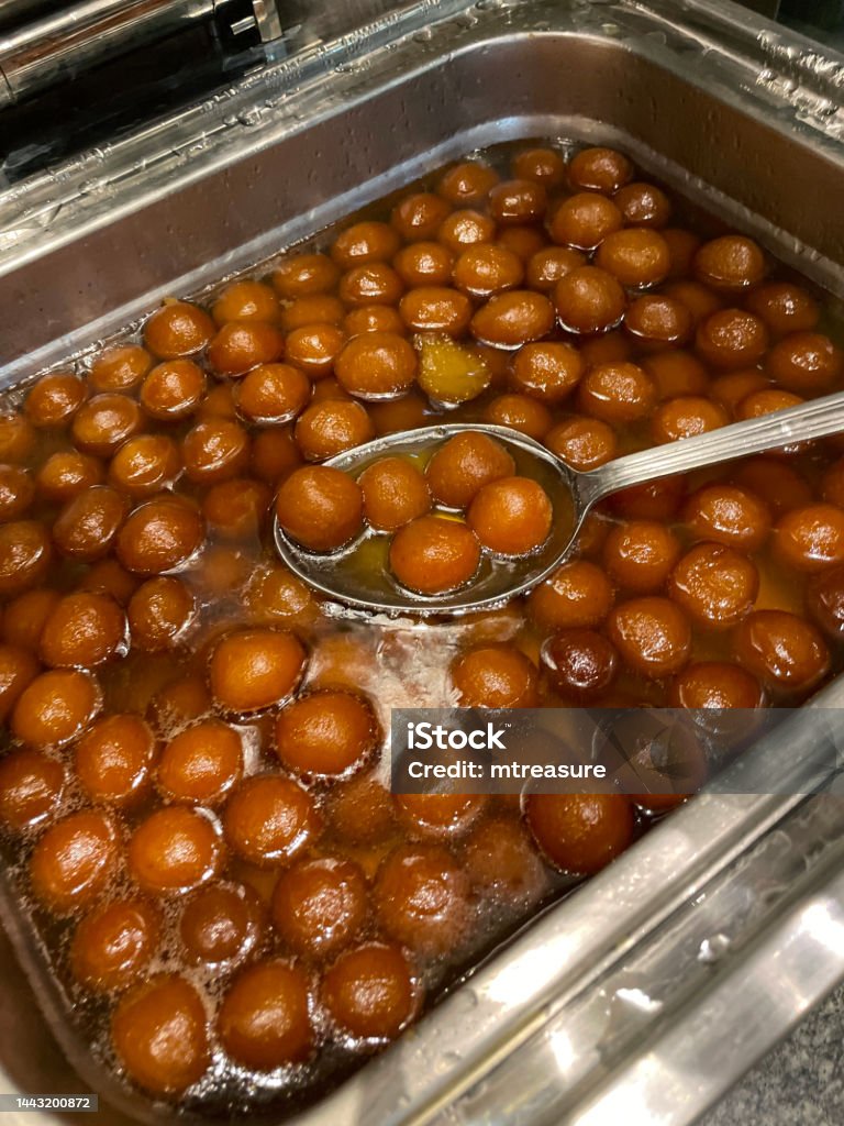 Image of catering tray of Gulab jamun (Rose water berry) Mithai (sweets) in Indian candy shop display, soaked in a sugar syrup (water, sugar cardamom, rosewater, and saffron), elevated view, focus on foreground Stock photo showing close-up view of tray containing Indian sweets in a sweet shop. These candies are the national dessert of India. The balls are called Gulab jamun and are made from khoya (dried evaporated milk solids) dough rolled, deep fried and then soaked in a sugar syrup (water, sugar cardamom, rosewater, and saffron). Cardamom Stock Photo