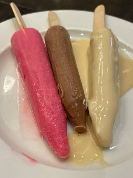 istock Full frame image of white side plate on a table containing Kulfi (frozen dairy dessert ) on wooden lolly sticks, strawberry, chocolate and vanilla flavoured ice cream custard, elevated view 1443203165