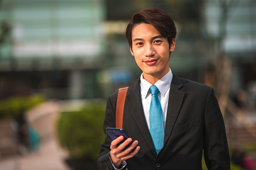 Outdoor portrait of handsome asian young man wearing a suit. He is looking at the camera with a smile and a cellphone on his hands.
