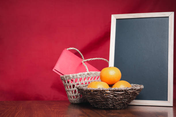 A small blackboard, four tangerines, and red envelopes in a basket on a wooden table with a red cloth background. Space for text. Happy Chinese New Year concept.