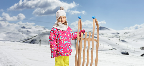 Child in winter clothes standing with a wooden sled on a snowy mountain