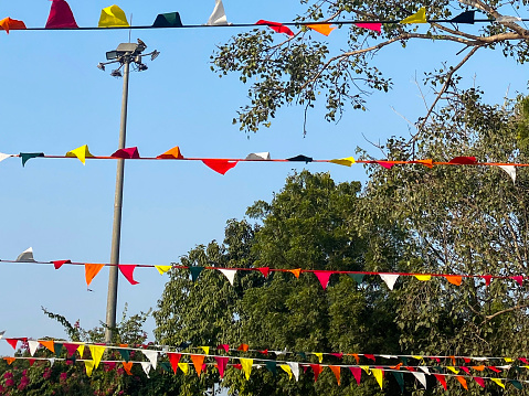 Photo showing some rows of triangular shaped bunting flags in orange, red, yellow, green and white, strung up at an outdoor event and pictured against the clear blue sky.