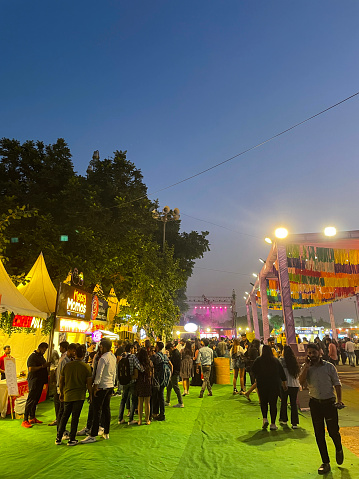 Horn Ok Please Food Festival, Delhi, India - November 11, 2022: Stock photo showing close-up view of groups of people wandering around exhibition grounds of Horn Ok Please Food Festival at dusk with illuminated lightbulb signage and fairground lights.