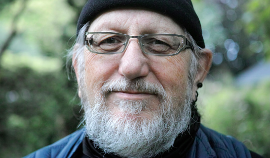Portrait of a 68-year-old senior man wearing blue clothes and a black cap. He has a beard, glasses and black earings. The background is blurred