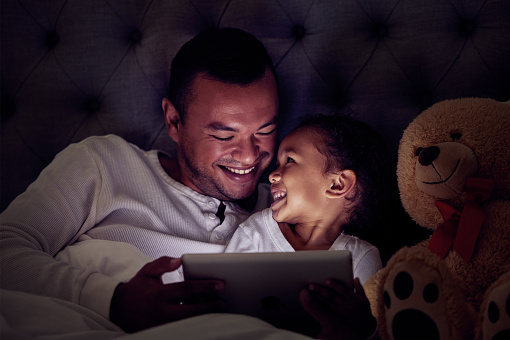 Father and child in bed with tablet reading ebook or to watch a film together at night. Happy girl with her dad enjoying online live streaming movie, game or digital kid app in dark home bedroom
