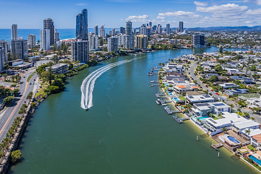 Aerial view Nerang River Housing estate (Isle of Capri) with Surfers Paradise and Pacific Ocean in the background. In the foreground a speedboat (private craft) creates a wake in the river.  Luxury waterfront houses with swimming pools, private boat moorings & piers.