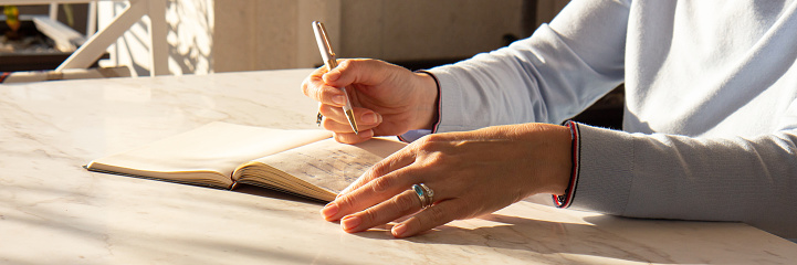 Stylish woman writing in a notebook placed on a bright desktop in the sun. Female makes notes in a notebook at home. Concept of freelancing, psychology, thinker, education. Banner