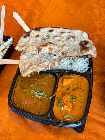 Stock photo showing elevated view of Indian takeaway meal in a black, plastic, single-use, disposable container divided in to sections, on an orange tablecloth. The meals consist of pilau rice, chicken tikka masala, chicken jalfrezi curry, and naan bread.