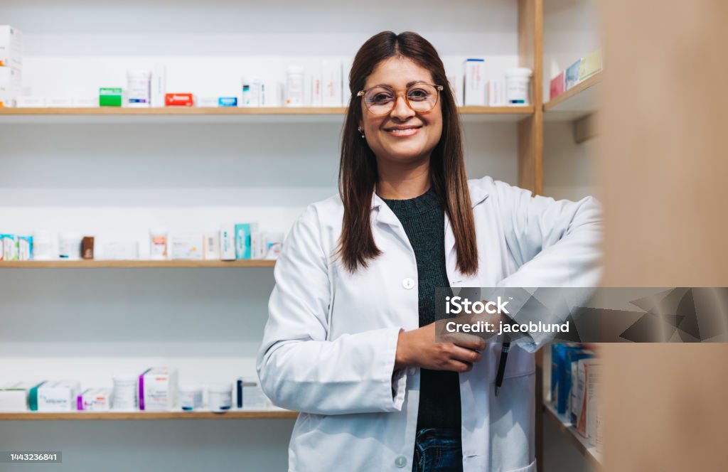 Female pharmacist standing in a drug store Female pharmacist standing in a drug store and looking at the camera. Happy woman working in a pharmacy. Portrait of a female healthcare worker in a chemist. Pharmacist Stock Photo