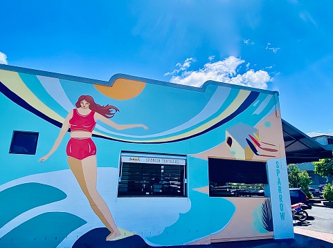 Horizontal side of public street community mural of woman surfing waves on blue concrete wall with blue cloudy sky above in Byron Bay Australia